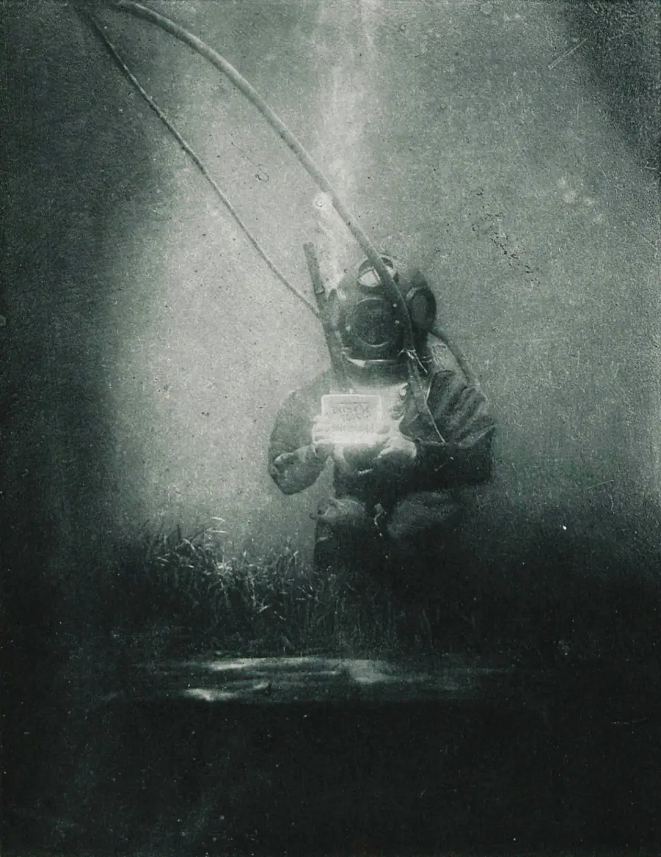 An 1899 underwater photograph: a diver in a hardhat suit standing on the seafloor holding a slate that reads Photographie Sous Marine.