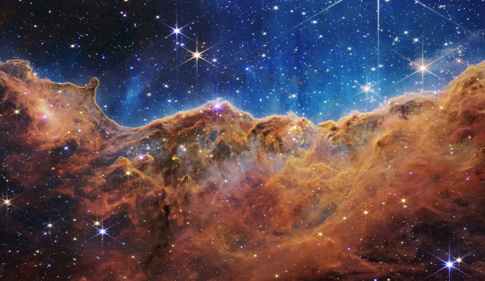 The Cosmic Cliffs of the Carina Nebula photographed by the James Webb Space Telescope: glowing amber mountains of gas under a deep blue starfield.
