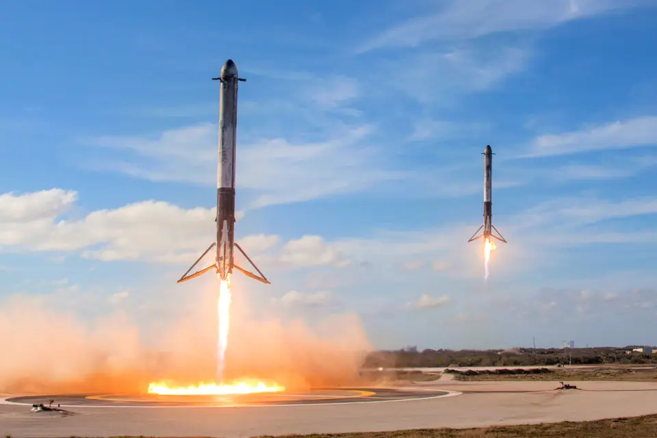 Two Falcon Heavy side boosters landing side by side at night on neighboring pads, twin pillars of flame under them.