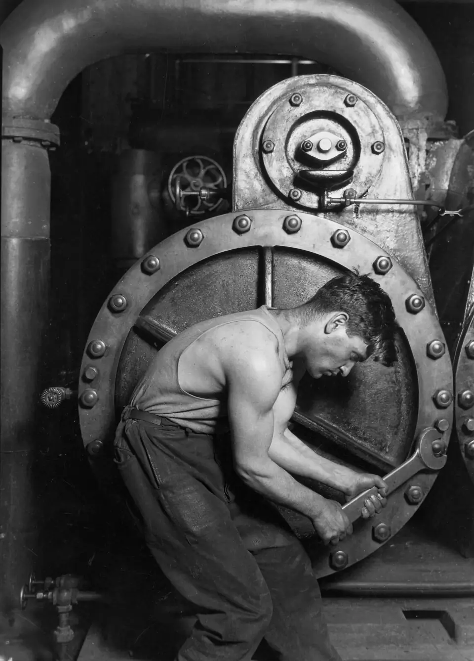Powerhouse mechanic, 1920: a muscular worker curled into the circle of a huge steam pump wheel, wrench in both hands.