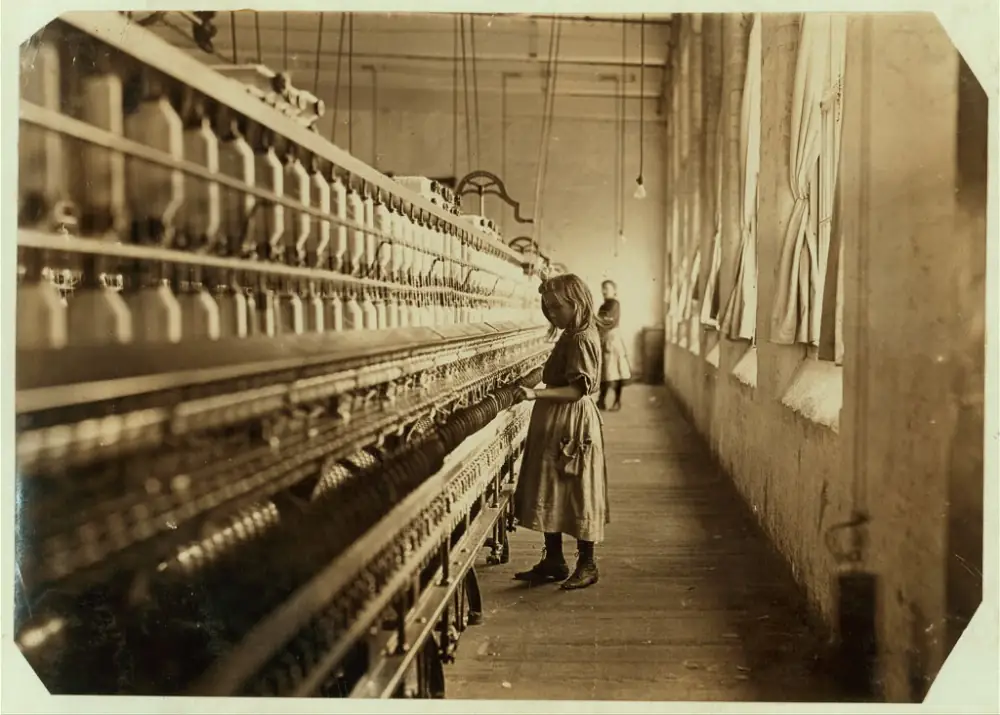 Sadie Pfeifer, a small girl in a work dress, standing at a towering cotton spinning frame in a 1908 South Carolina mill.