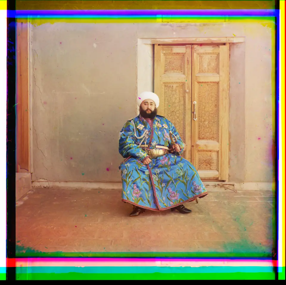 The Emir of Bukhara in 1911, in full color: a bearded man in a brilliant blue silk robe with a gold belt, rainbow fringes at the photo's edges.