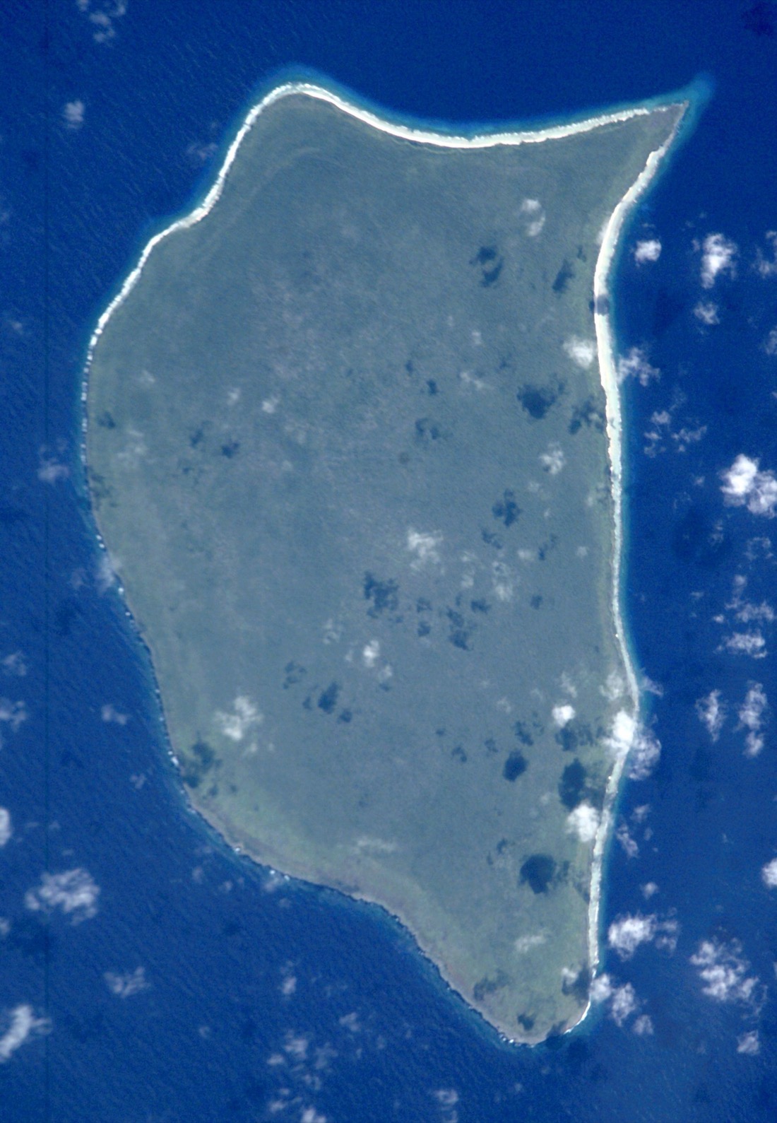 A small island ringed by pale reef and dark ocean, seen from high above in space.