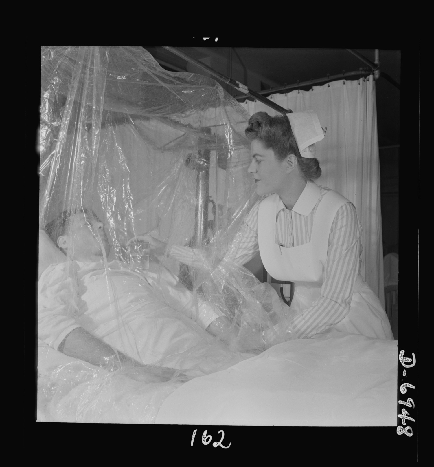 A nurse at the bedside of a man lying under a clear oxygen tent in a 1942 hospital room.