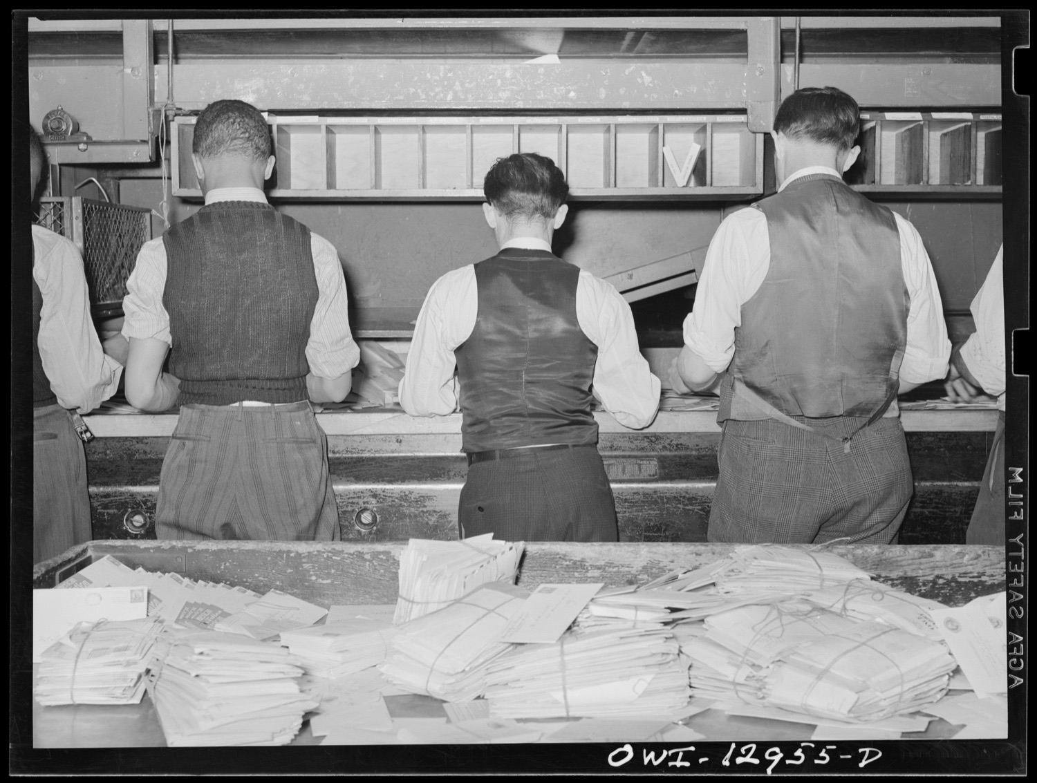 Clerks at sorting cases behind heaps of tied bundles of letters in a 1938 mail room.