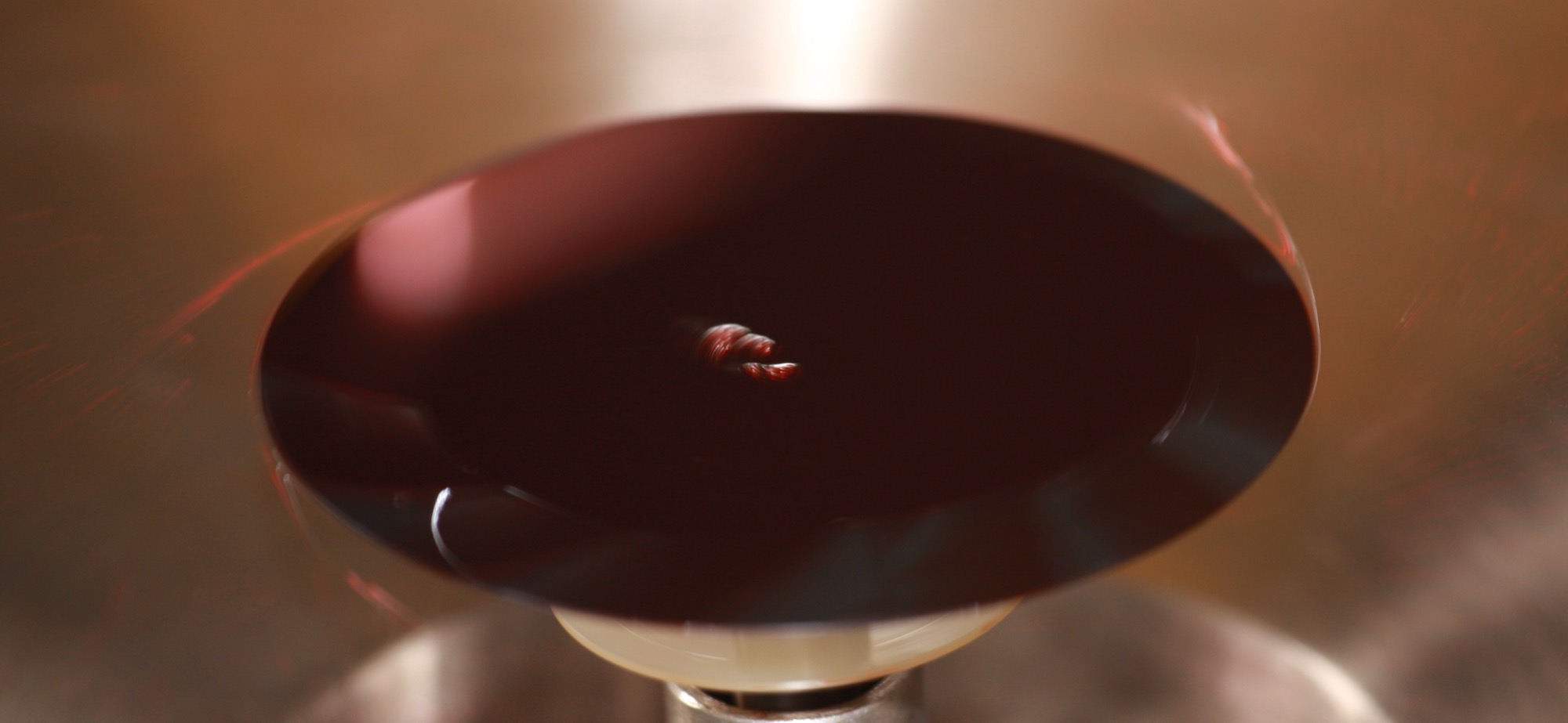A silicon wafer spinning under a coat of deep red photoresist, the liquid flung into glossy ripples and streaks by the spin.