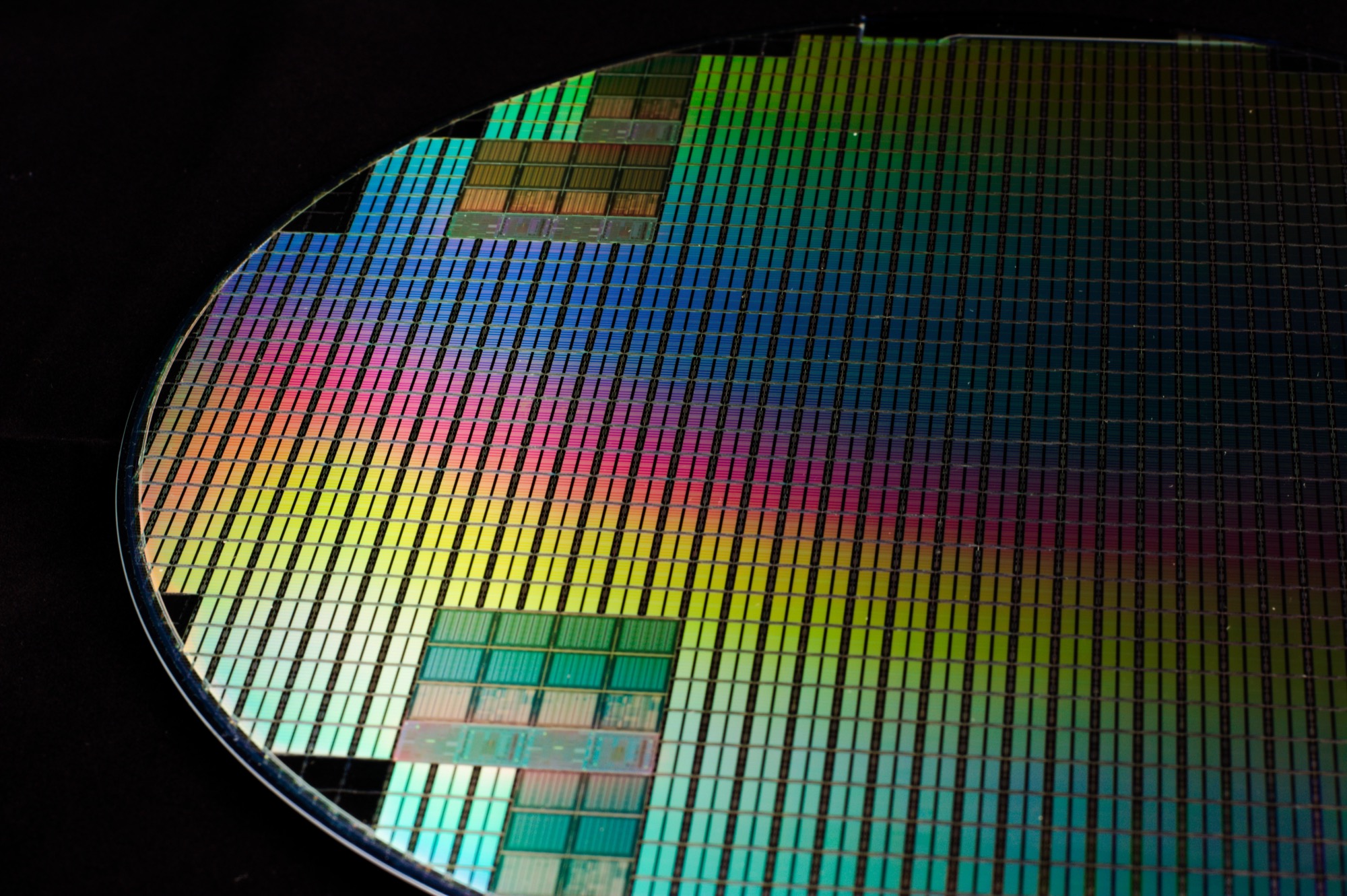 A patterned silicon wafer at a raking angle on black, its grid of identical chips breaking the light into a vivid rainbow.