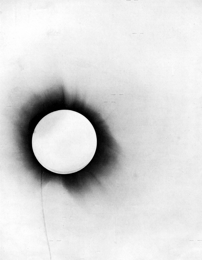 The 1919 total solar eclipse photographic negative, a dark disk of the eclipsed Sun ringed by faint corona, with short horizontal marks identifying the positions of stars near the Sun.