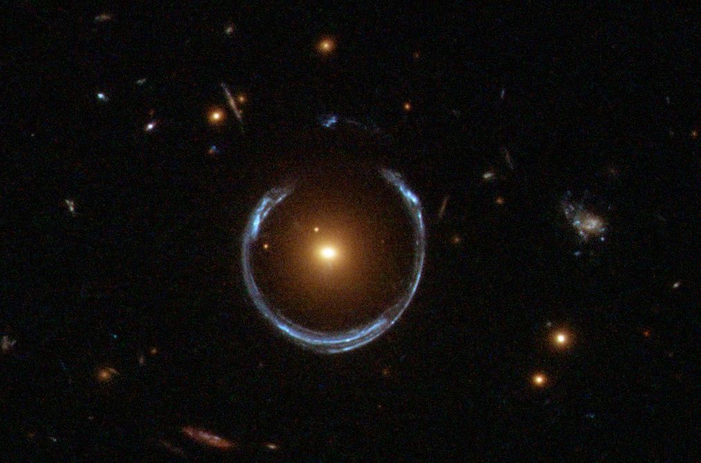 A Hubble image of the Cosmic Horseshoe: a bright blue near-complete ring of light wrapped around a glowing red galaxy at its center.