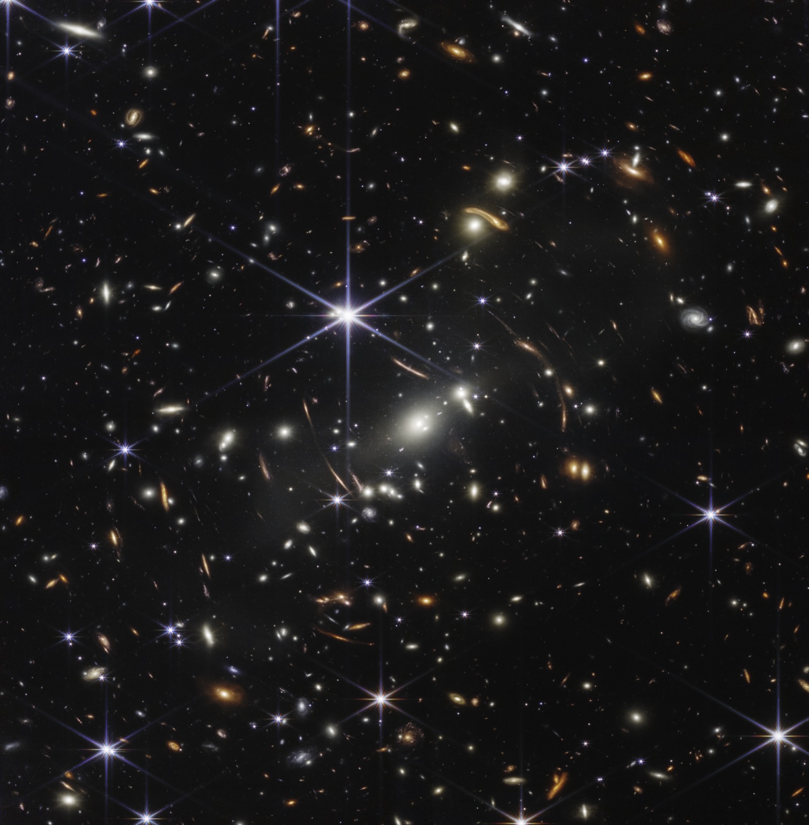 The James Webb Space Telescope's First Deep Field: thousands of galaxies of every shape and color scattered across a black sky, some smeared into curved arcs by gravitational lensing.