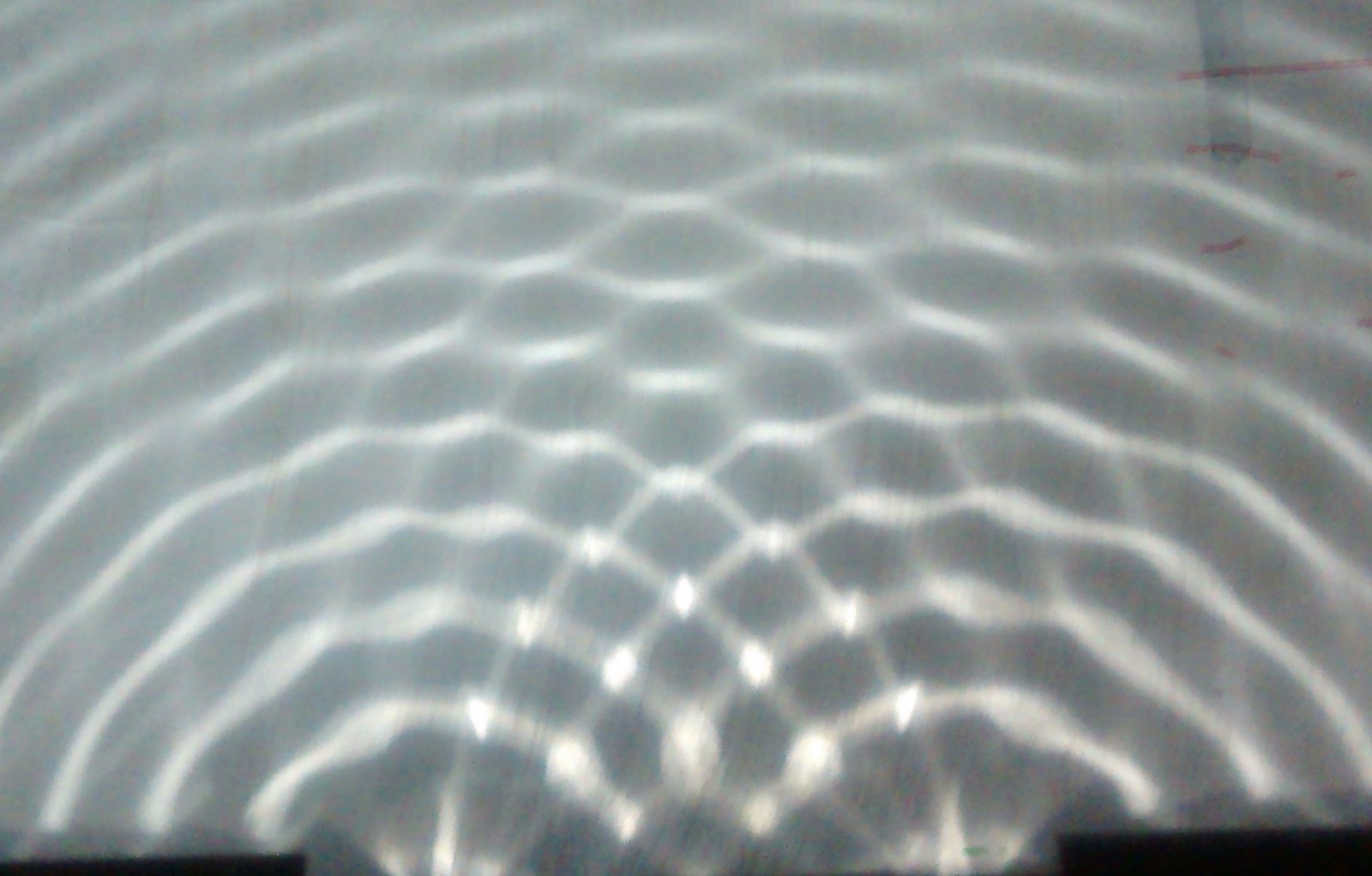 A ripple tank photograph: two sets of circular water waves spreading from two points and overlapping into a pattern of bright and dark fan-shaped bands.