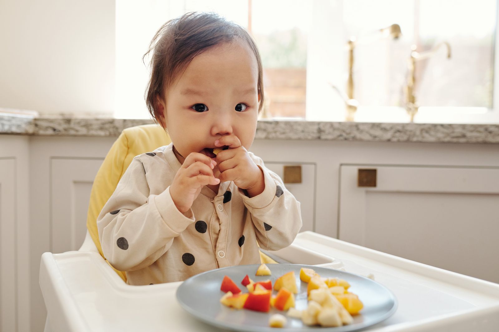 A baby in a high chair picking up soft pieces of fruit from a plate.