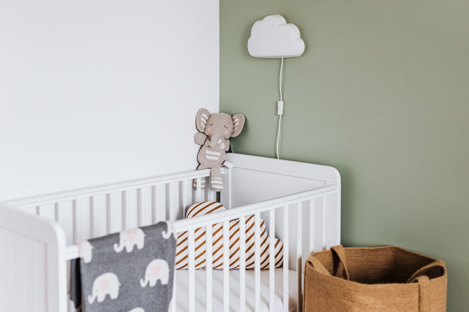 A calm nursery corner: a white crib against a sage-green wall, a cloud-shaped lamp, a soft elephant toy, and a woven basket.
