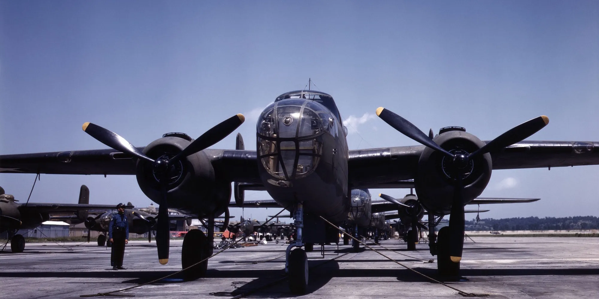 Rows of olive-drab B-25 bombers on an outdoor assembly line under a deep blue sky, with a single worker standing among them, Kansas City, 1942.