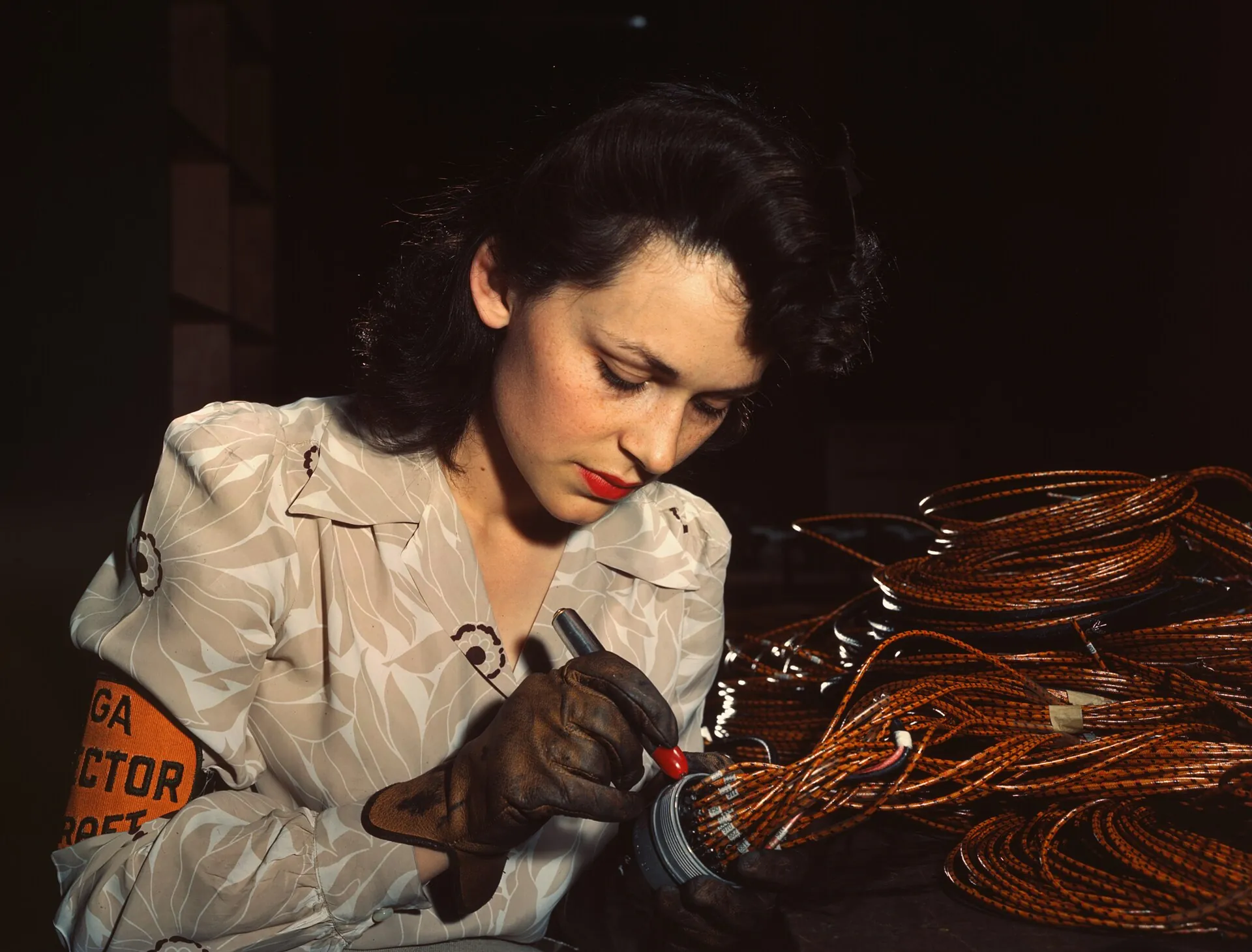 A worker in a red headscarf and plaid shirt checking rows of electrical assemblies at a workbench, in saturated 1942 Kodachrome color.