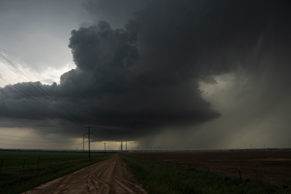 A supercell thunderstorm towering over a dirt road through flat farmland.