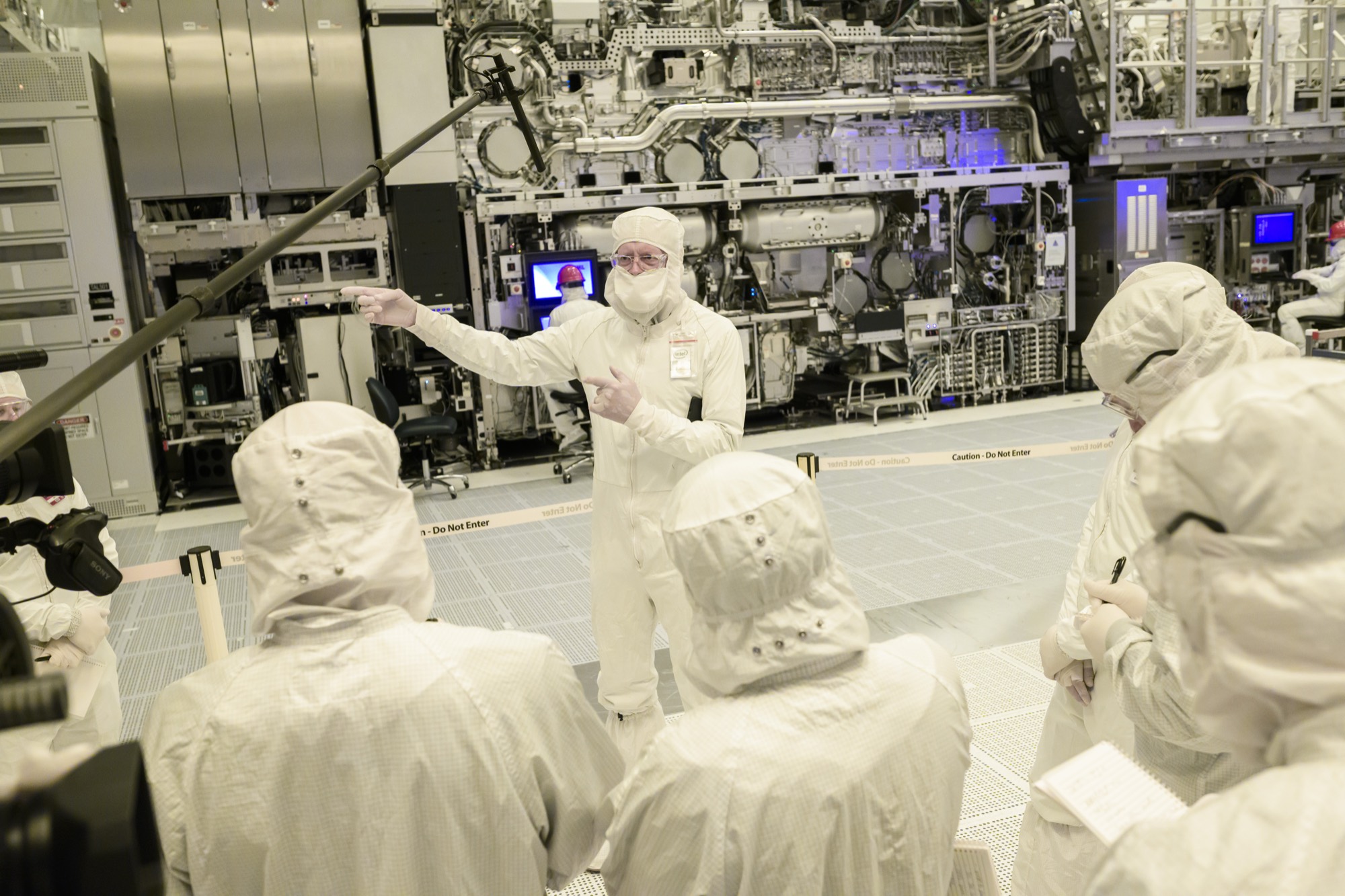 People in white cleanroom bunny suits being briefed in front of the lithography machine, one pointing.