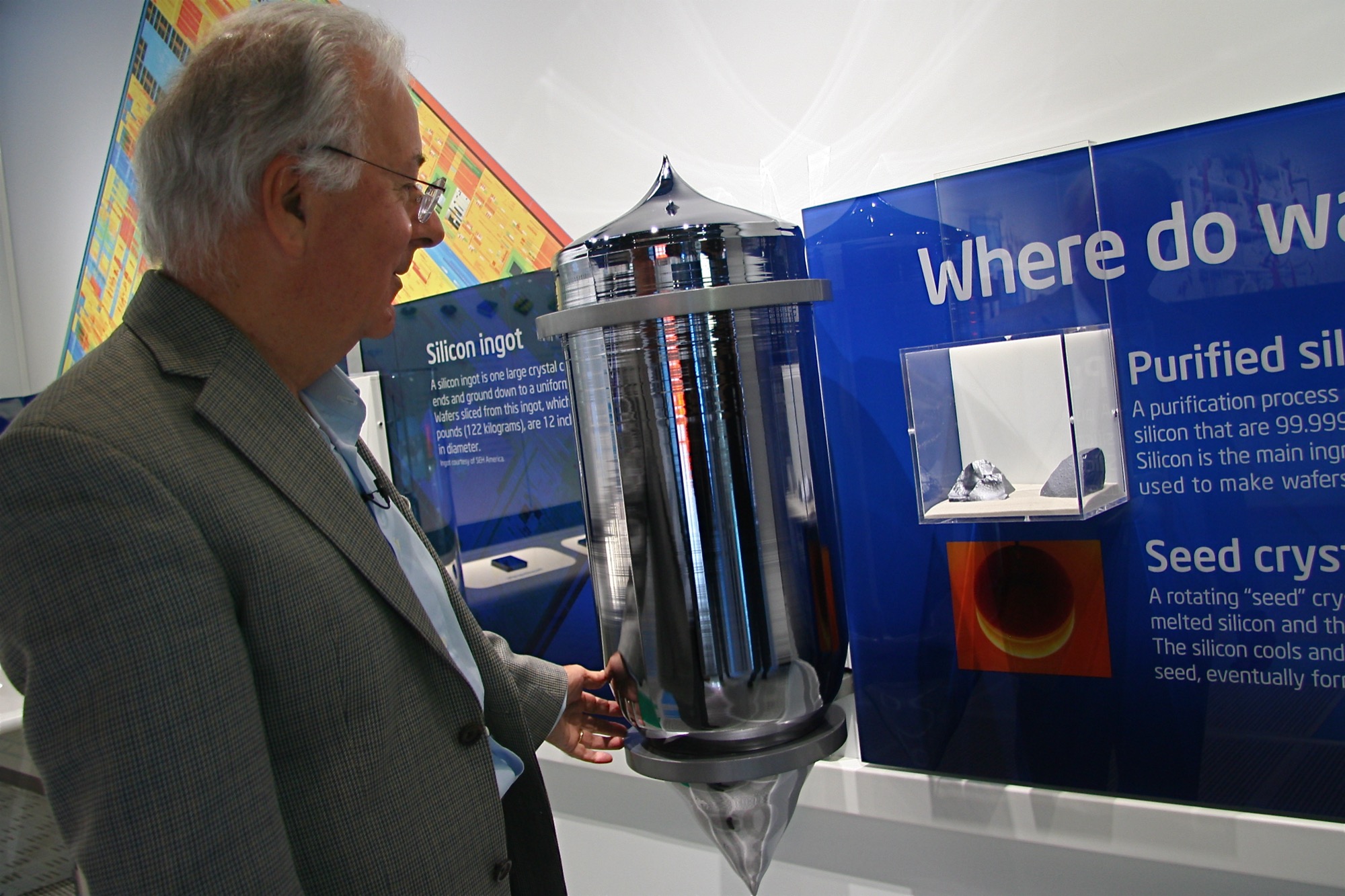 A man in a jacket standing beside a tall mirror-polished cylindrical silicon ingot in a display case; the ingot rises from his waist to above his shoulder.