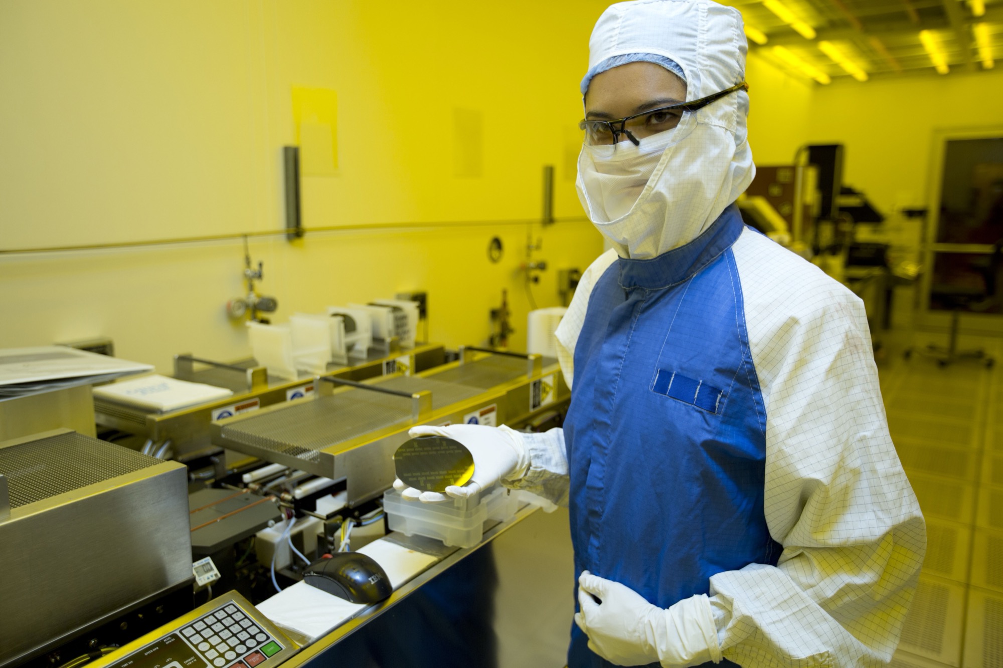 A worker in a yellow cleanroom, in mask and gloves, holding a blank dark mirror-finish silicon wafer.