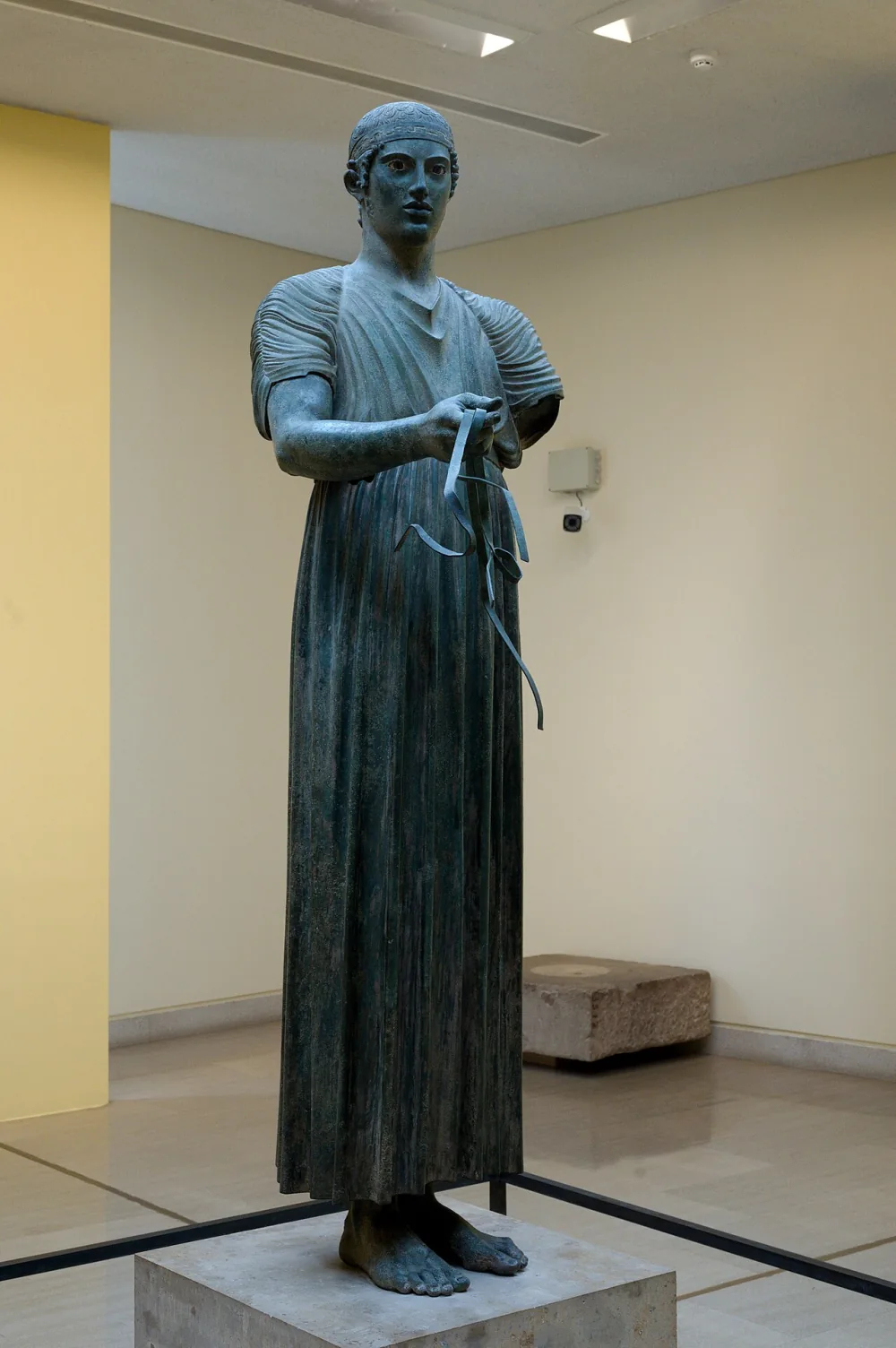 The Charioteer of Delphi, a life-size ancient Greek bronze of a young charioteer in a long pleated robe, standing calm and upright with one arm extended to hold the reins.