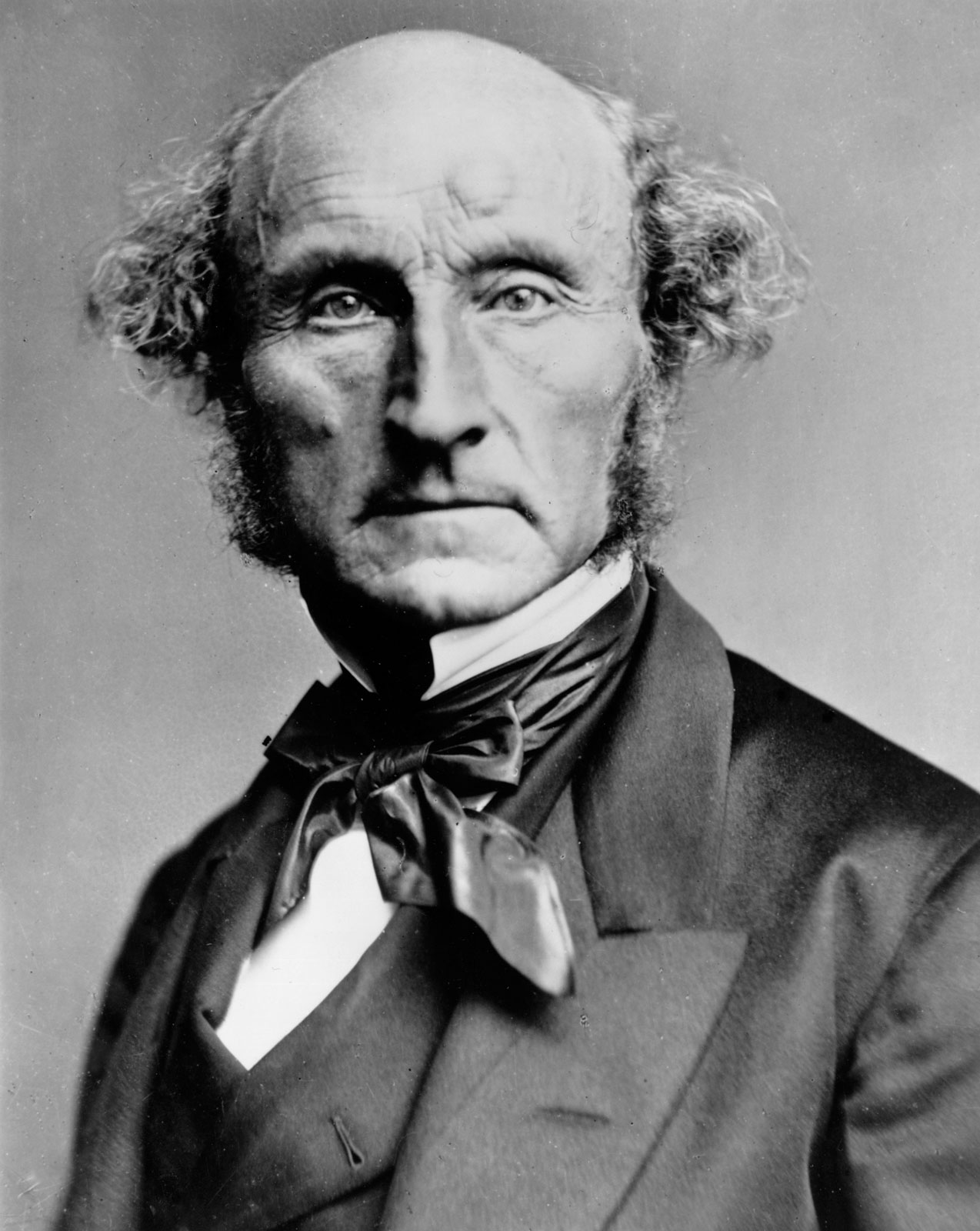 A c.1870 photograph of John Stuart Mill: a bald, aging man with curling side hair and long sideburns, in a dark coat and silk cravat, looking straight at the camera with a grave, deeply lined face.