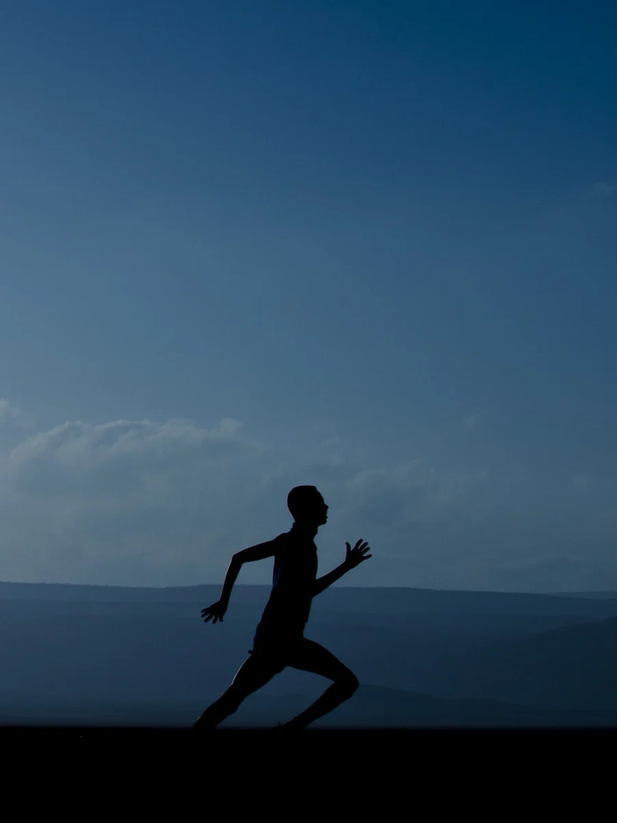 A lone runner in silhouette, mid-stride, against a vast blue dawn sky and distant desert mountains.
