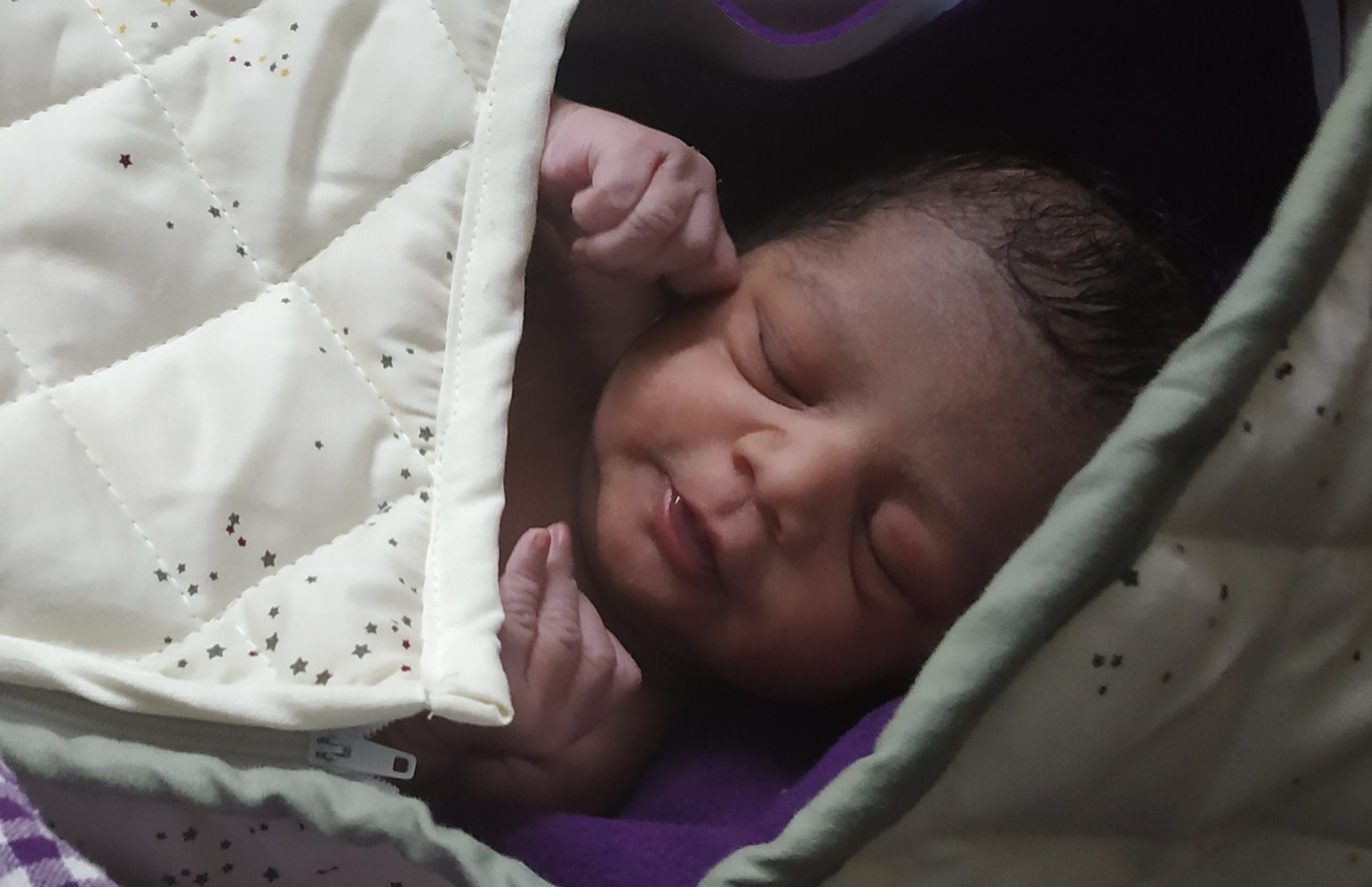 A sleeping newborn on its first day, wrapped in a pale star-print blanket, both tiny fists curled up beside its face.