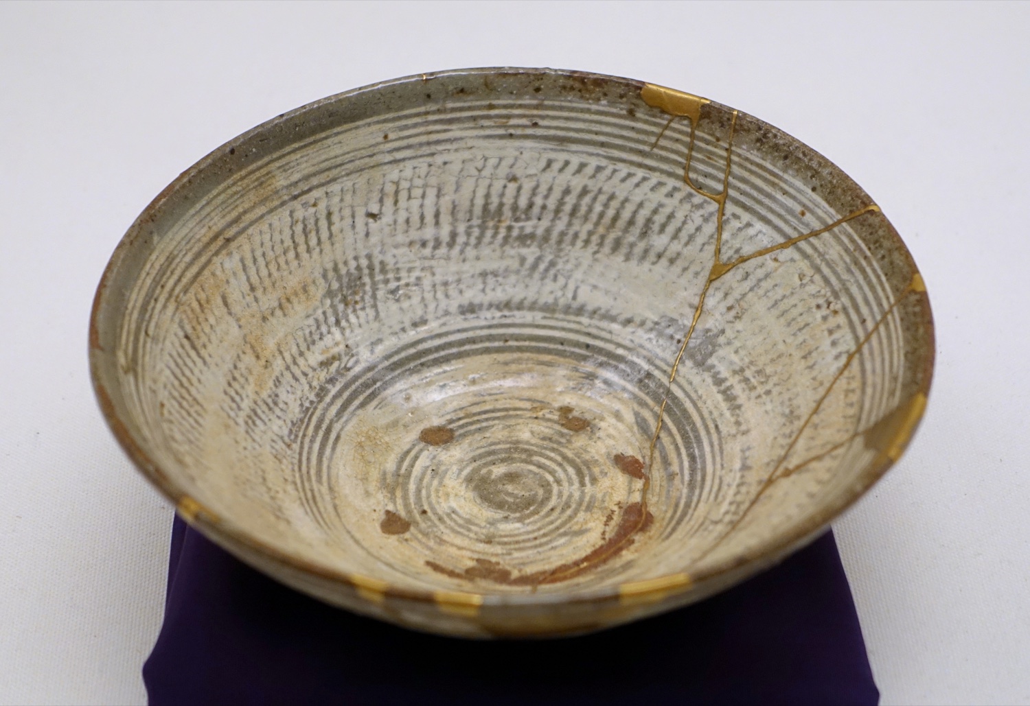 A pale 16th-century Korean tea bowl repaired in the kintsugi method, its cracks filled and traced with bright seams of gold lacquer.
