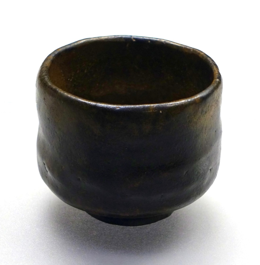 A black, thick-walled, slightly irregular Raku tea bowl on a plain ground.