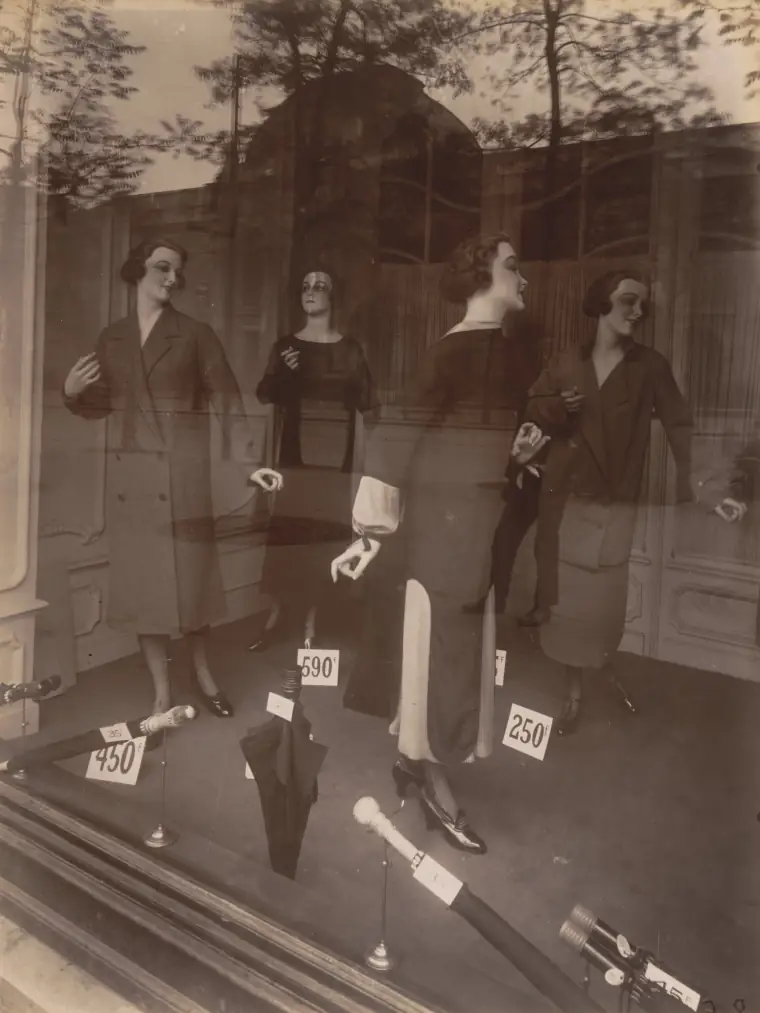 A 1925 Atget photograph of a Paris shop window: suited mannequins glowing behind glass, the street reflected over them.