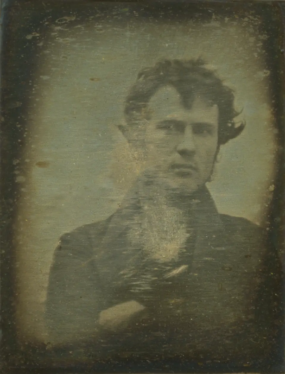 Robert Cornelius in 1839: a young man with windblown hair and crossed arms, looking warily at the camera, slightly off-center.