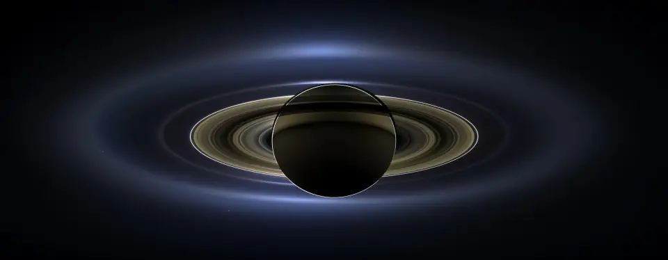 Saturn backlit by the Sun, its rings glowing, with Earth visible as a tiny pale blue dot below the rings.