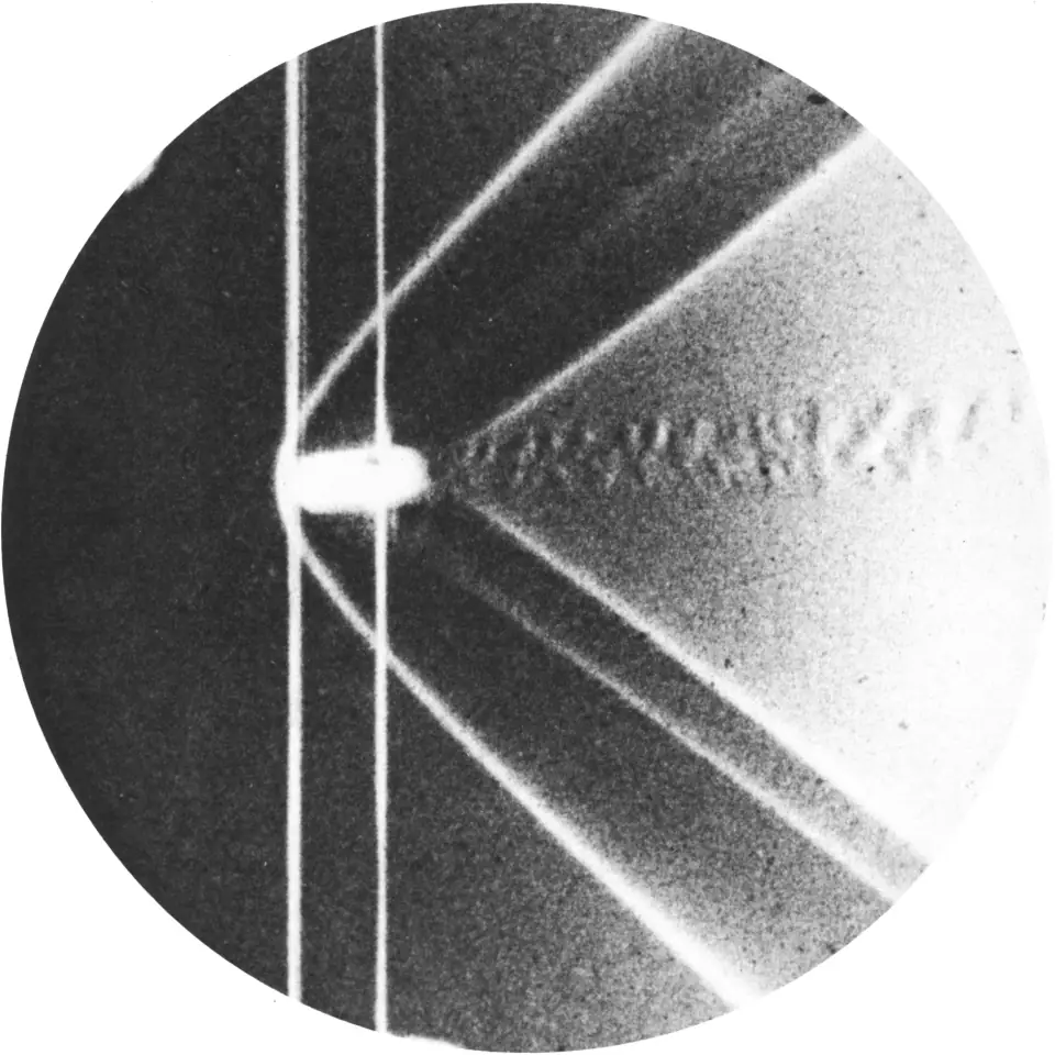 An 1888 shadowgraph of a supersonic bullet: the dark projectile trailing a visible cone-shaped shock wave through the air.
