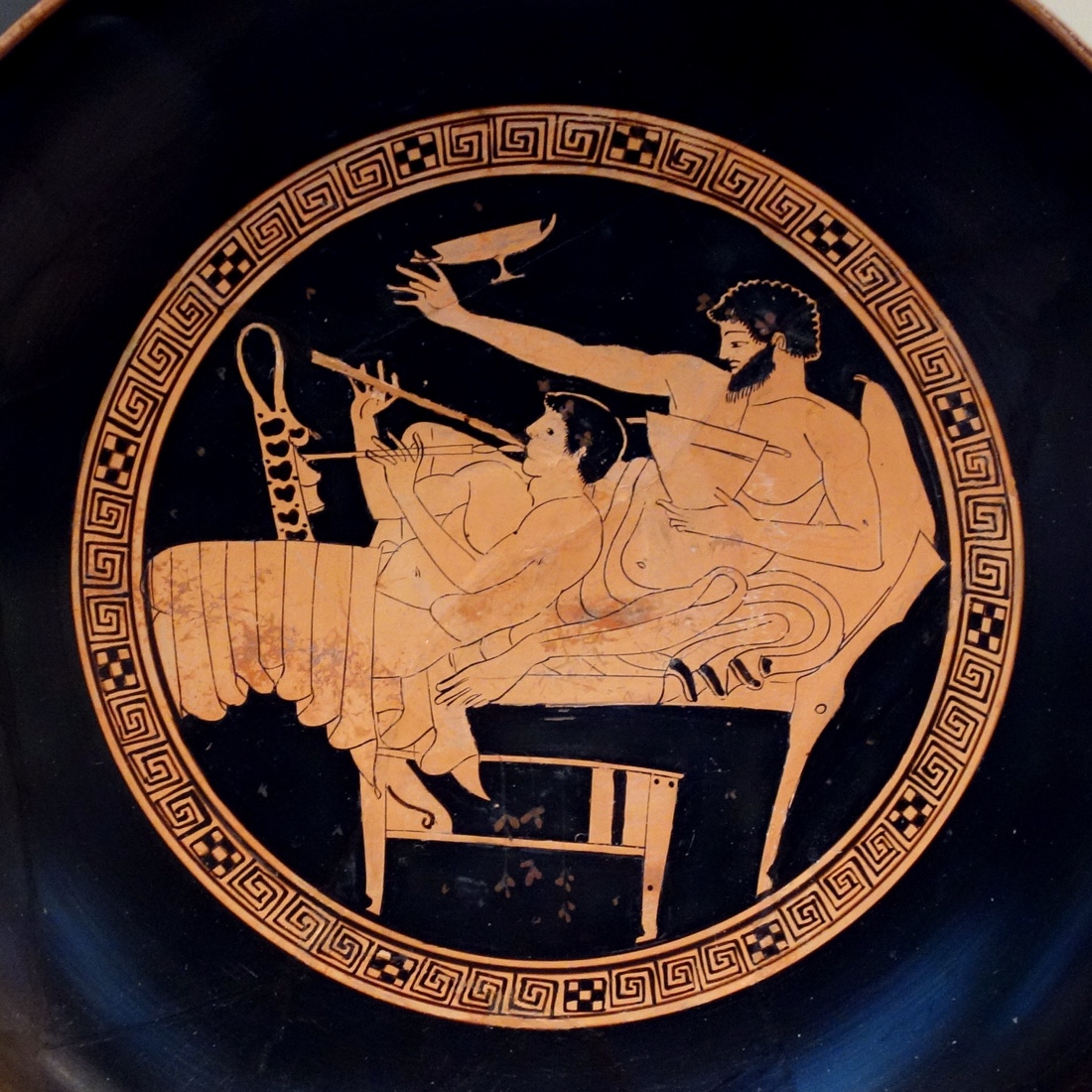 The painted interior of an ancient Greek red-figure drinking cup: a bearded man reclines on a couch raising a shallow wine cup while a youth beside him plays the double pipes, all framed by a Greek-key border.