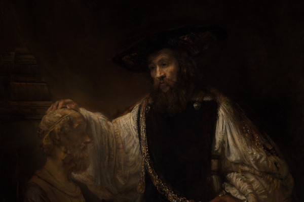 Rembrandt's painting of a bearded Aristotle in a black hat, lit gold against deep shadow, a heavy gold chain across his chest, resting one hand on a pale marble bust of Homer.