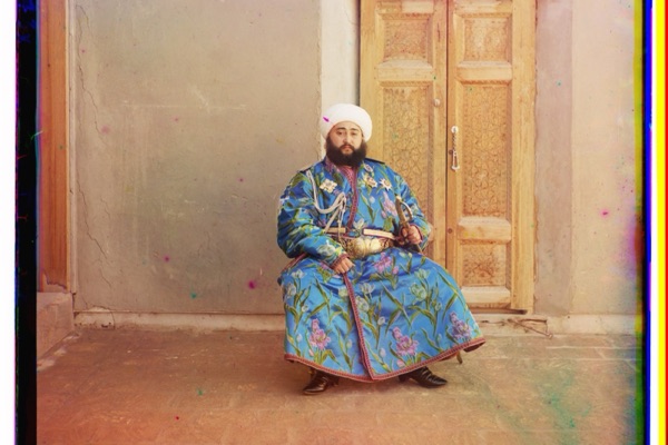 The Emir of Bukhara photographed in full color in 1911: a bearded man in a brilliant blue silk robe seated before a carved wooden door.