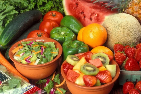 A market display crowded with tomatoes, peppers, cucumbers, melon, watermelon, strawberries, salad, and cut fruit.