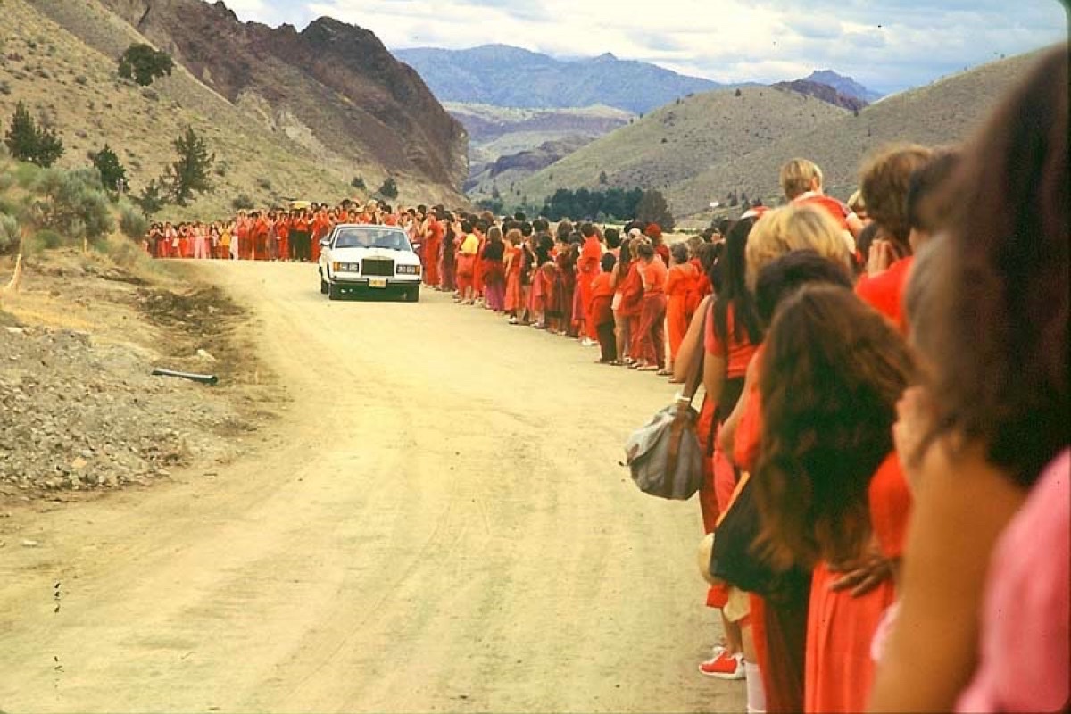 A white Rolls-Royce drives down a dirt road in the Oregon high desert, walled on both sides by hundreds of followers dressed in red, waiting to watch it pass.