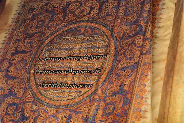 An illuminated page of the Guru Granth Sahib: Gurmukhi script in a central oval medallion, framed by ornate orange, blue, and gold floral borders on aged parchment.