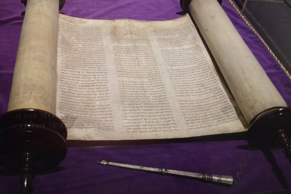 An open Torah scroll on purple velvet: three columns of handwritten Hebrew on parchment between two wooden rollers, with a silver pointer resting in front.