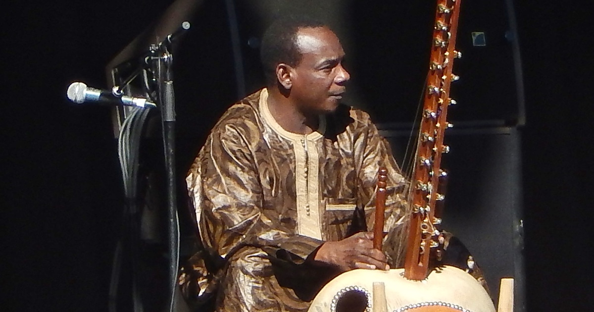 The Malian kora master Toumani Diabaté seated in profile, playing the kora, a tall harp with a large gourd body, against a dark stage.